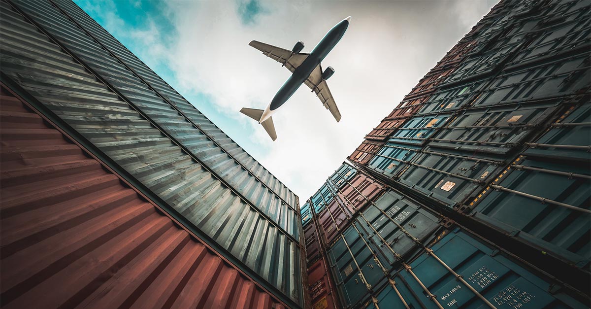Low-flying airplane passes over stacks of international shipping containers against an industrial backdrop