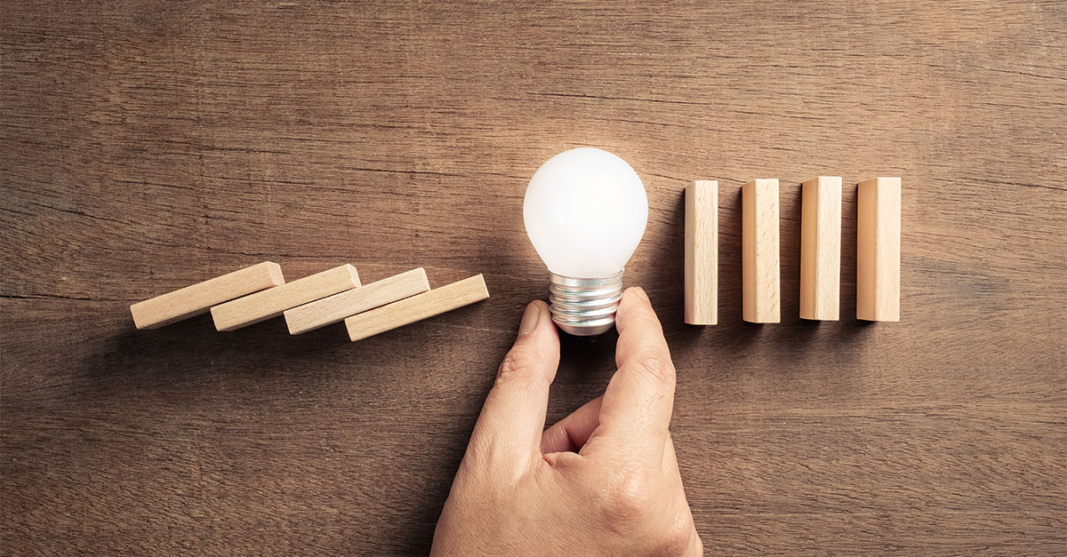 Hand stopping falling dominoes with a light bulb, representing proactive healthcare compliance, risk management, and operational strategy